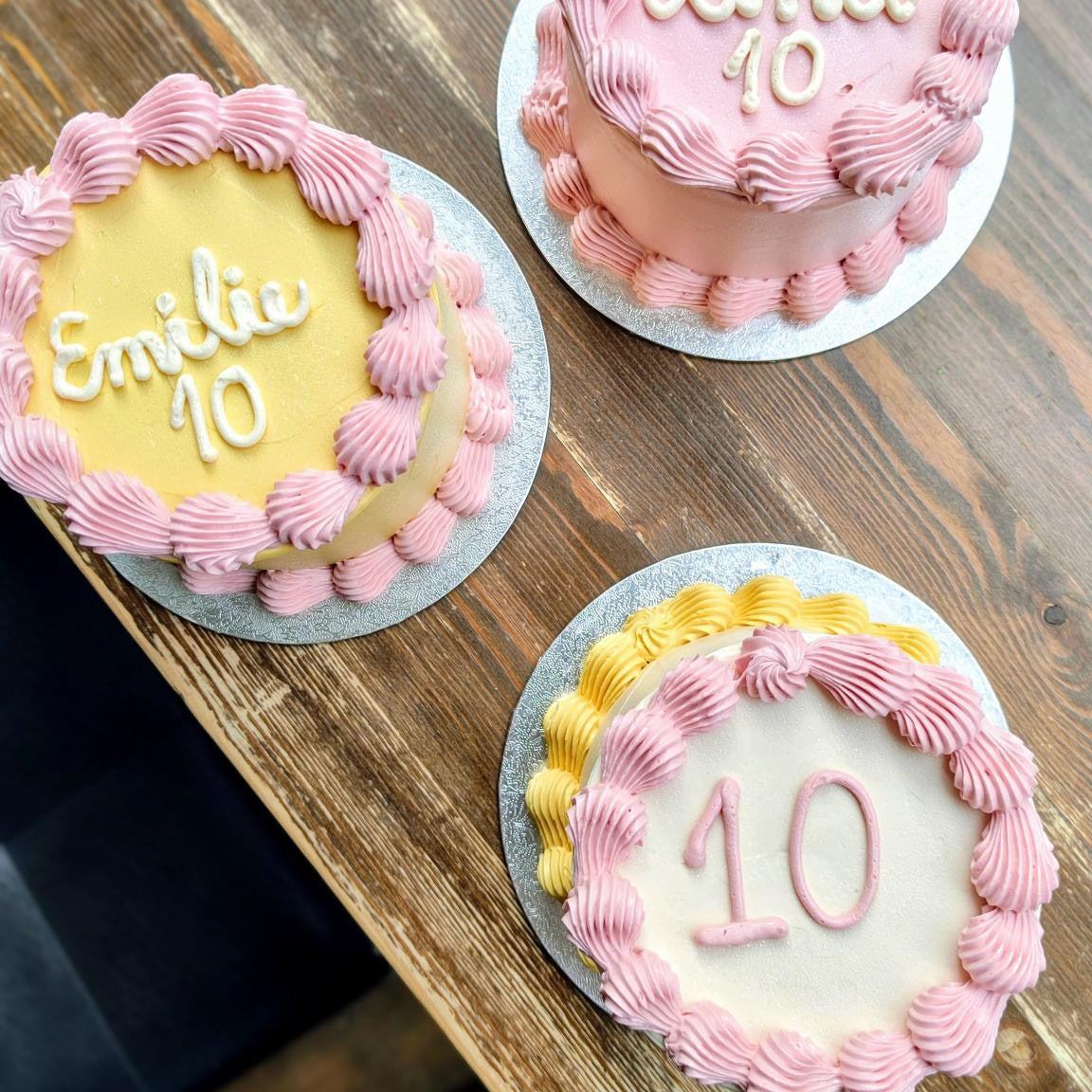 Three birthday cakes with pink and yellow icing on a wooden surface