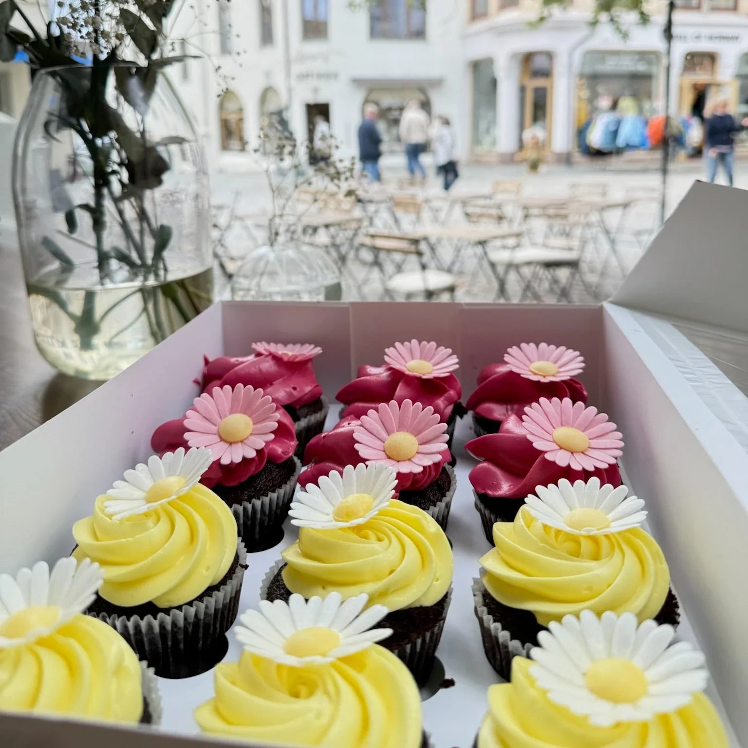 Box of cupcakes with yellow and white frosting and pink flowers, set against a blurred outdoor cafe background.