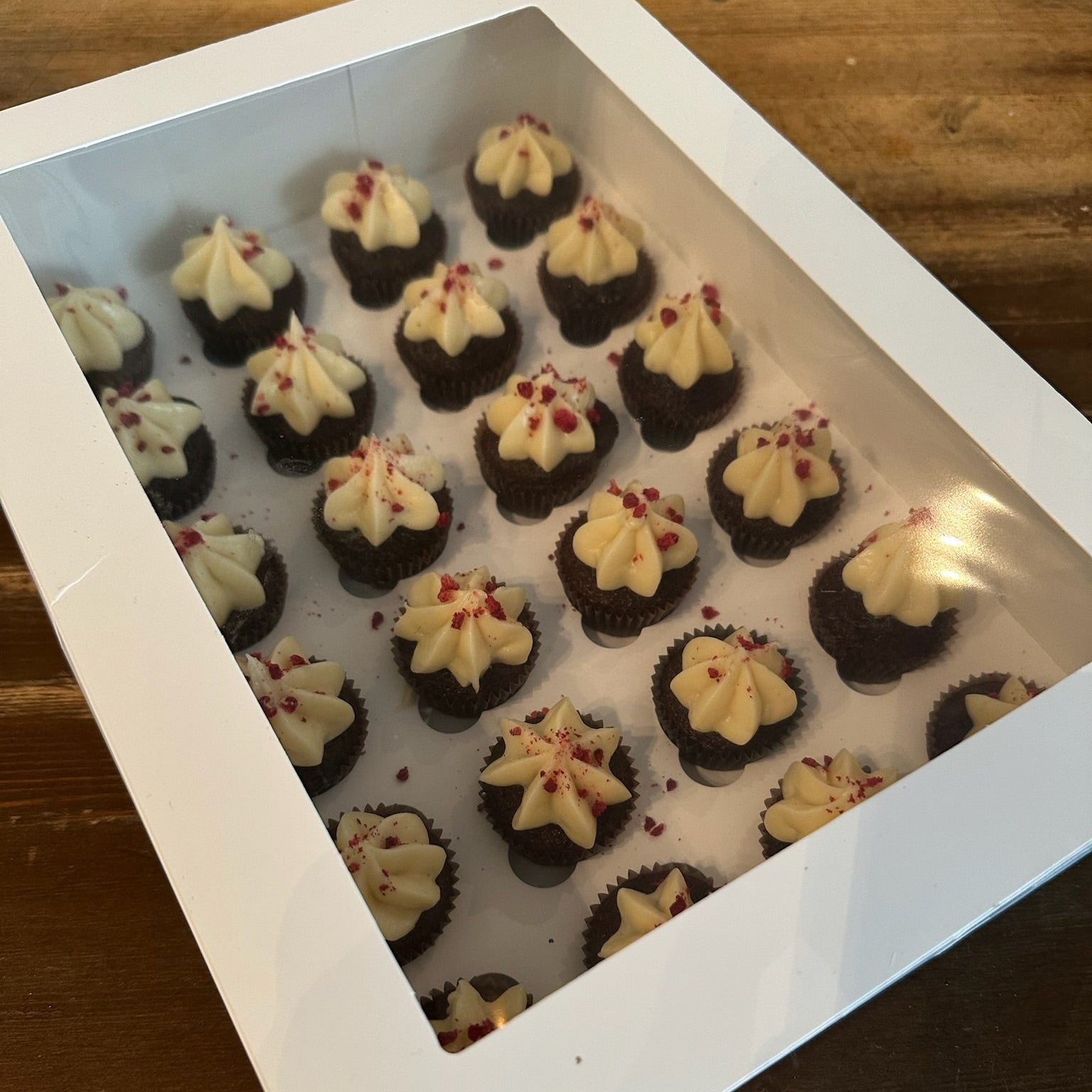 Mini chocolate cupcakes with white frosting and red sprinkles arranged in a box.