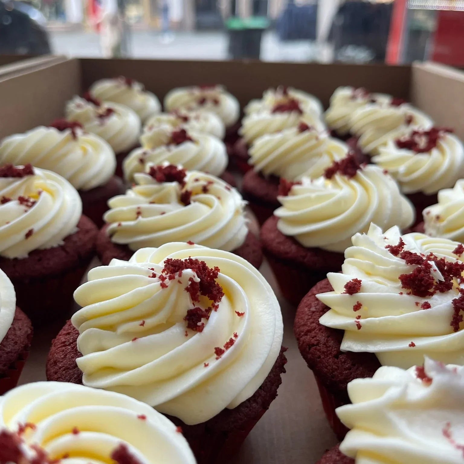 Red velvet cupcakes with white frosting and red velvet crumbs in a box.