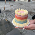 Hand holding a mini small rainbow-colored cake with white frosting on a cobblestone street.