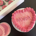 Heart-shaped pink cake with 'NORA' on top, surrounded by other similar cakes.