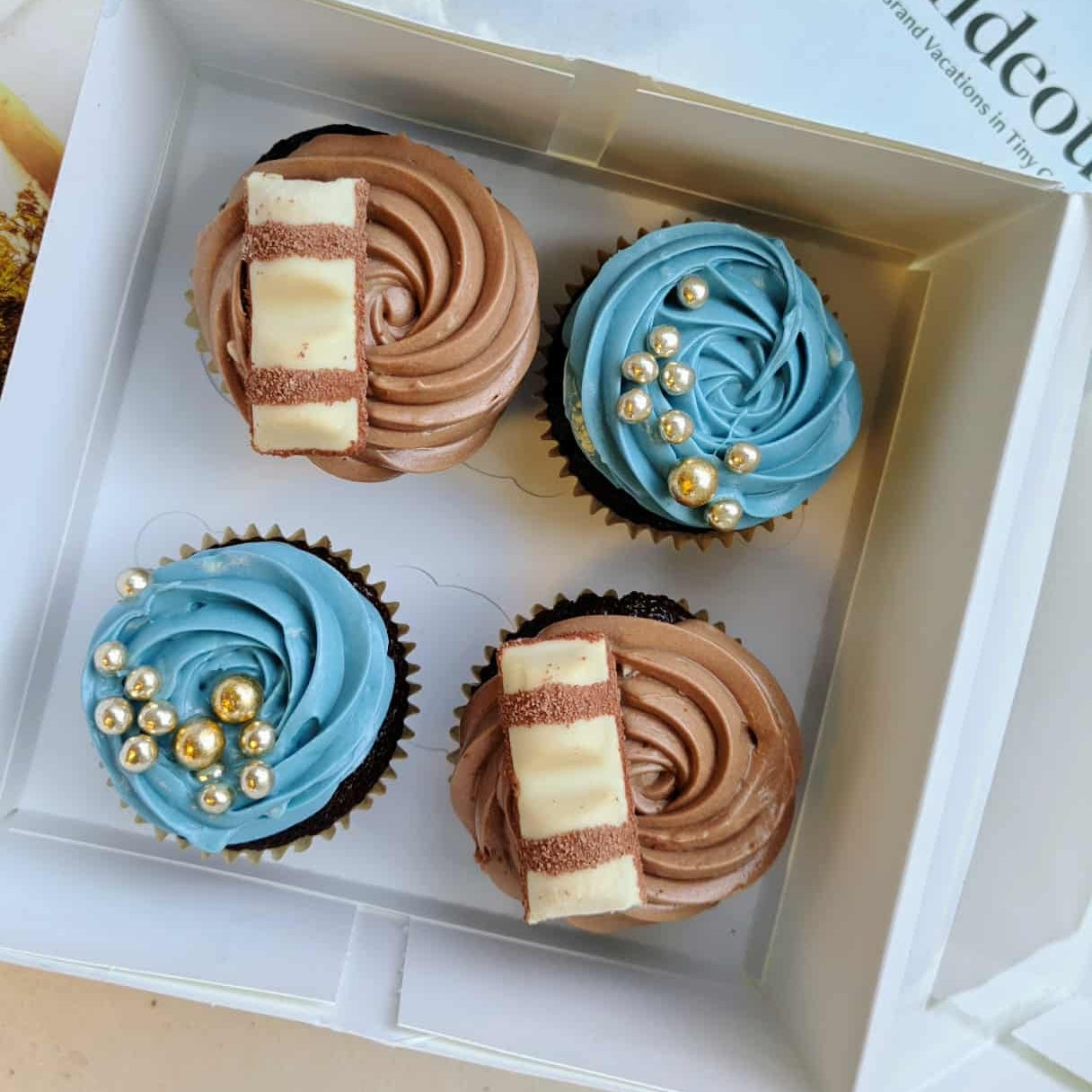 Four cupcakes in a box with brown, blue, and gold decorations.