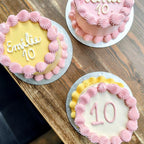 Three birthday cakes with pink and yellow icing on a wooden surface
