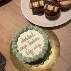 Small cake with text on a gold stand and plate of cupcakes on a wooden surface