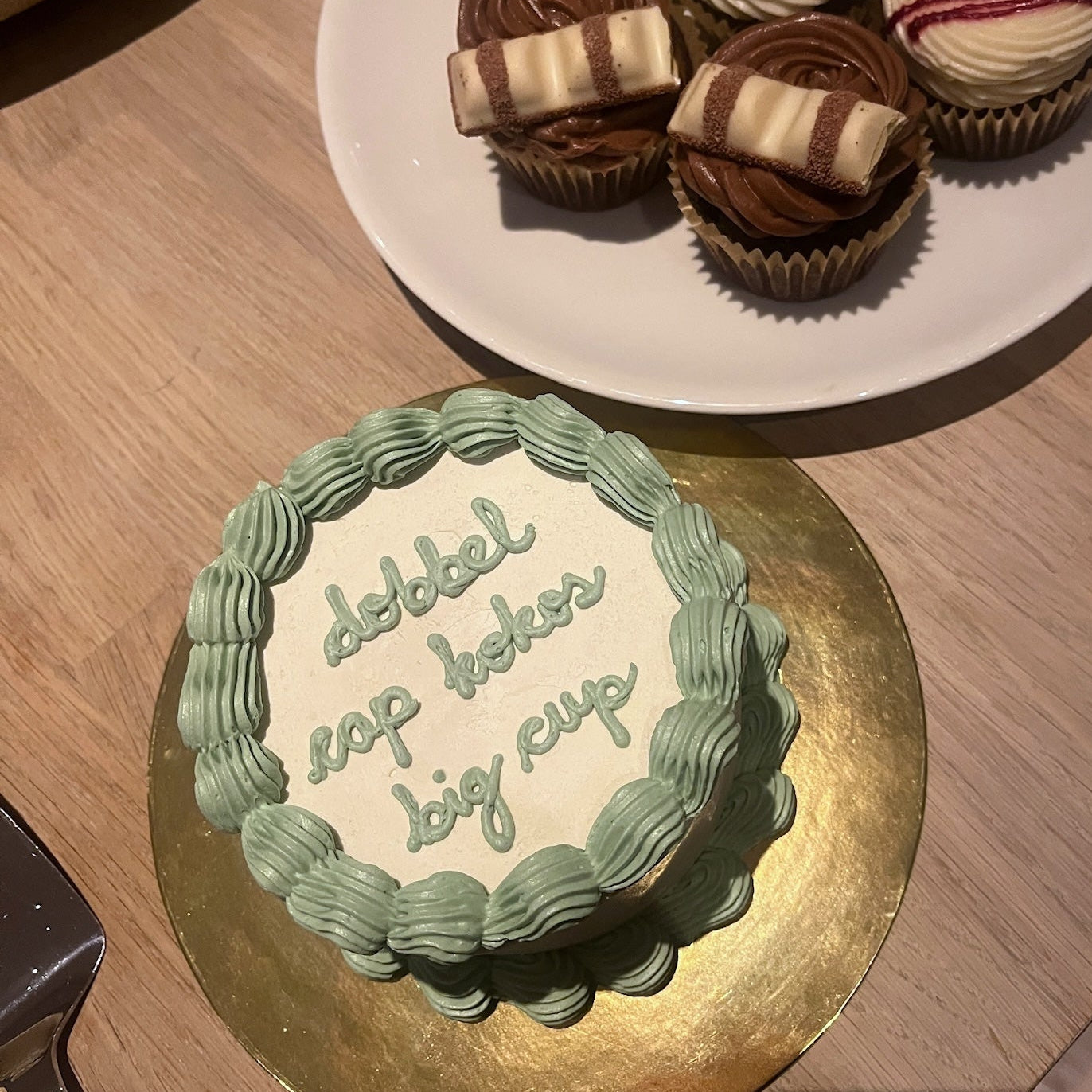 Small cake with text on a gold stand and plate of cupcakes on a wooden surface