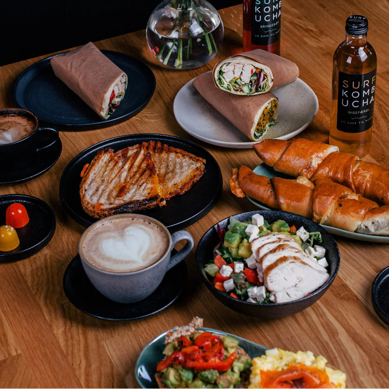 Wooden table with various food items including wraps, sandwiches, and a cappuccino, with a vase of flowers and bottles in the background.