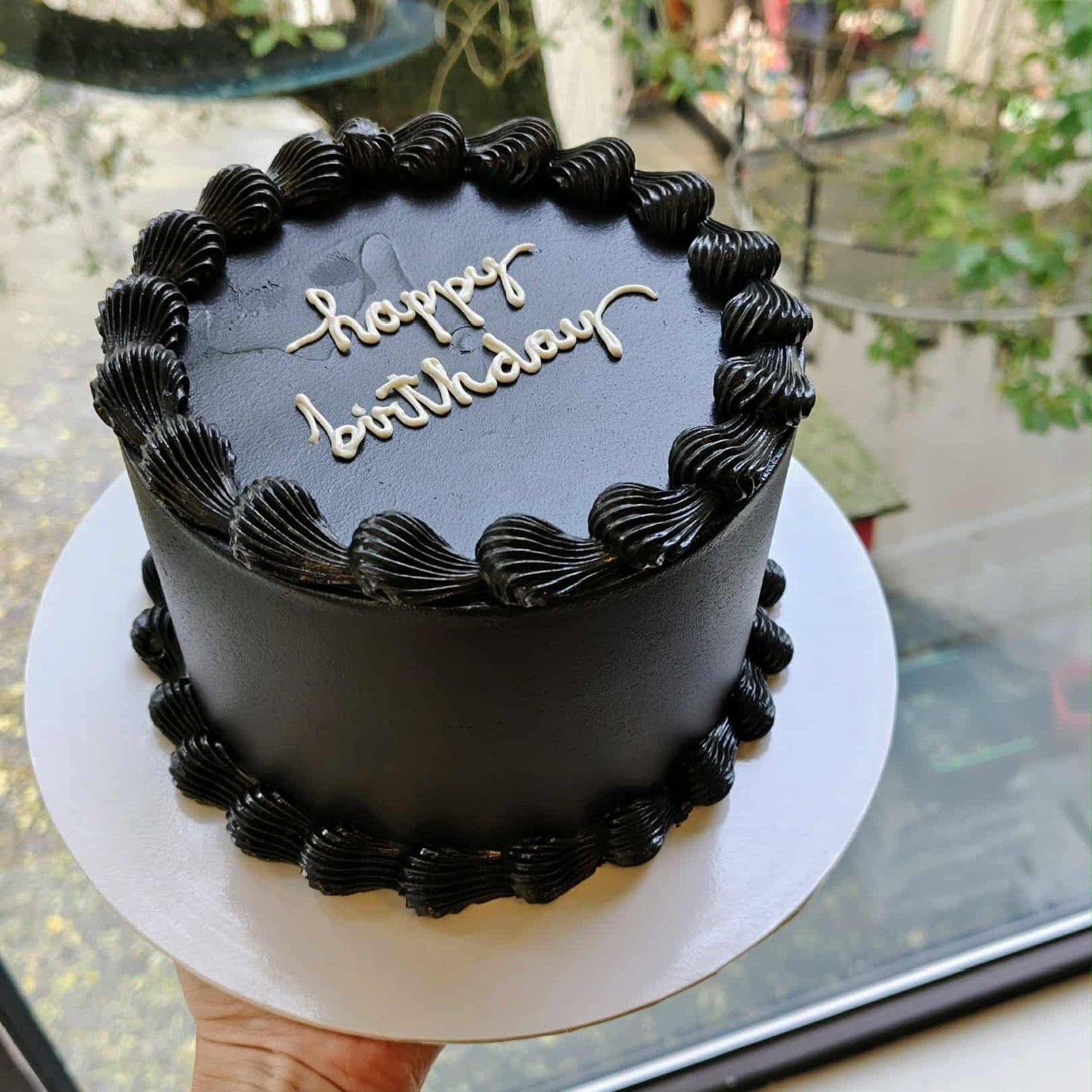 Black birthday cake with 'happy birthday' text held by a person indoors.